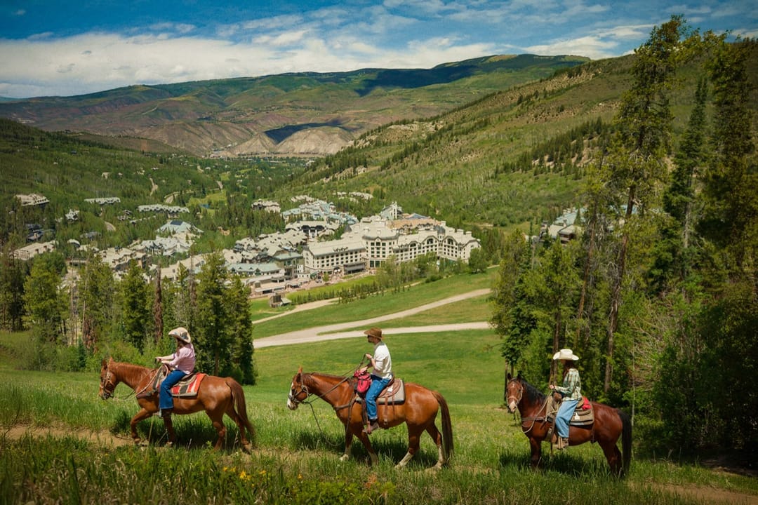 Three riders on horseback in a green meadow above the valley, Park Hyatt Beaver Creek in the distance
