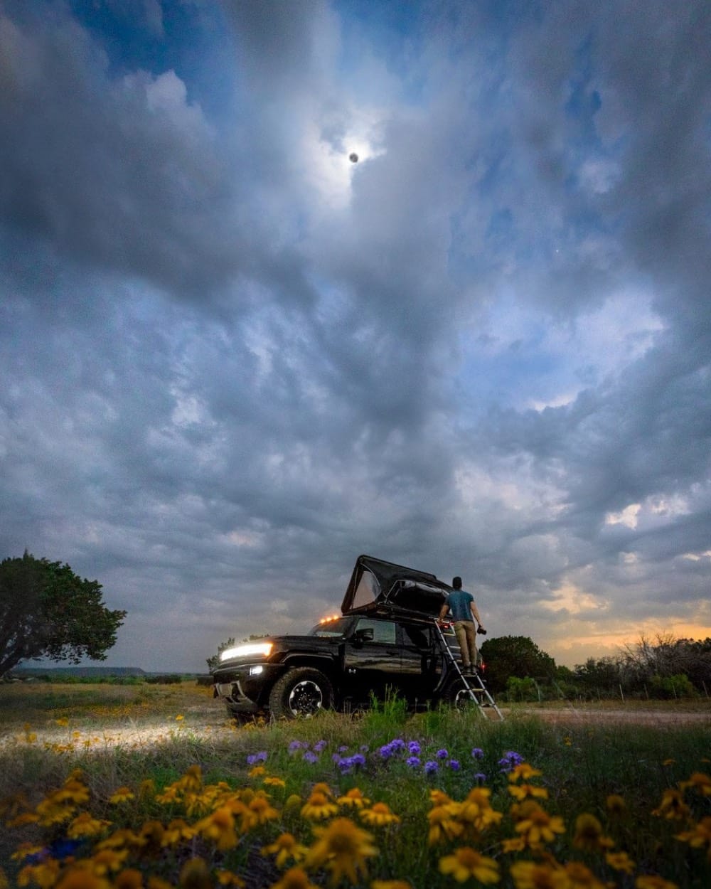 GMC Hummer EV with a rooftop tent under the 2024 total solar eclipse in Texas, a man on the ladder, wildflowers in front