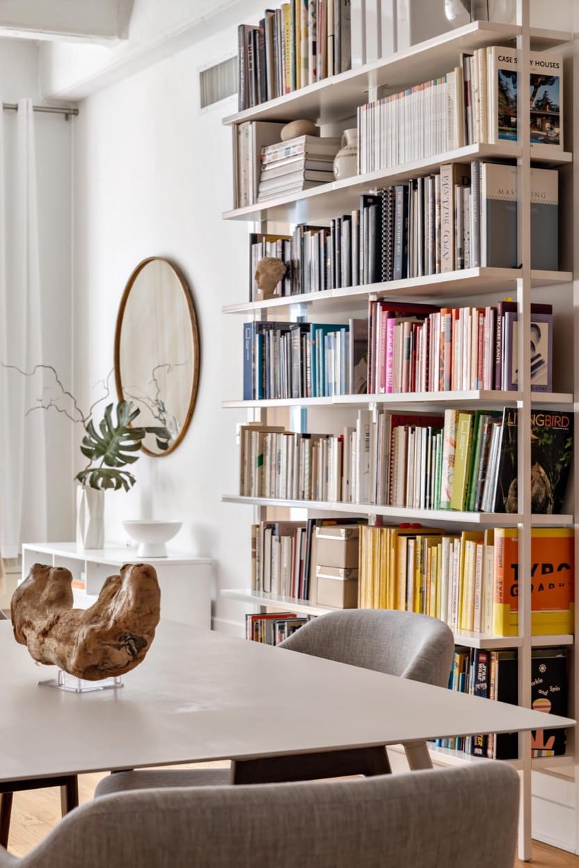 Dining table beside floor-to-ceiling bookshelves, a round mirror and a wooden sculpture