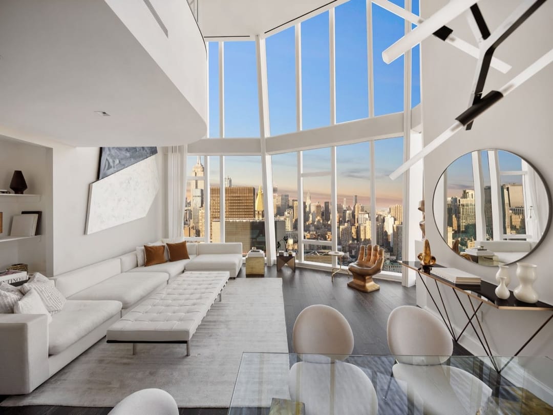 Double-height living room with white sofas and floor-to-ceiling windows onto Manhattan