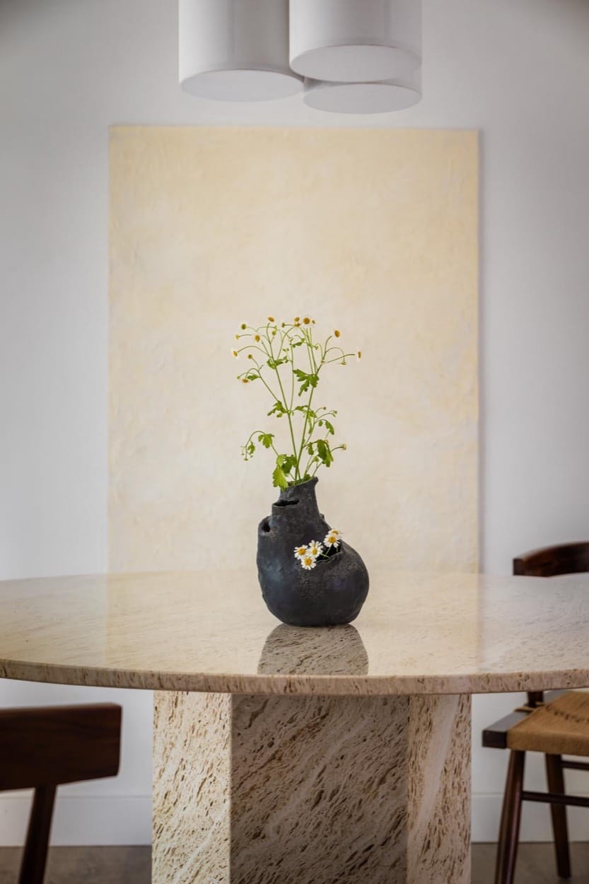 Stone pedestal dining table with a black vase of white flowers, a pale canvas behind