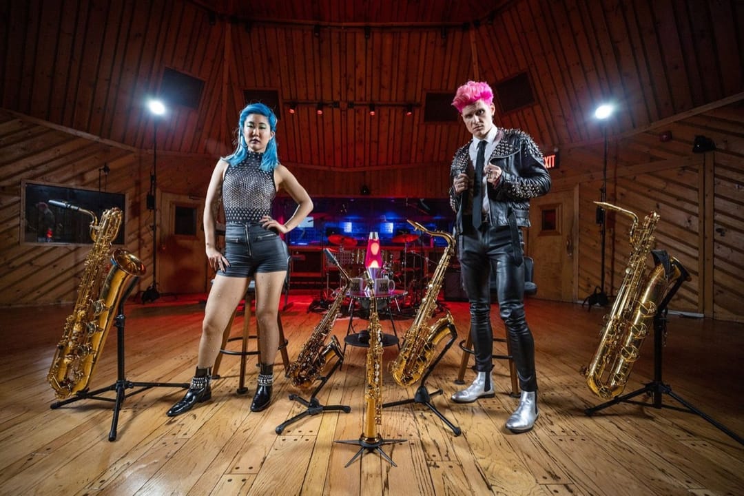 Two musicians with blue and pink hair among saxophones in a wood-paneled recording studio