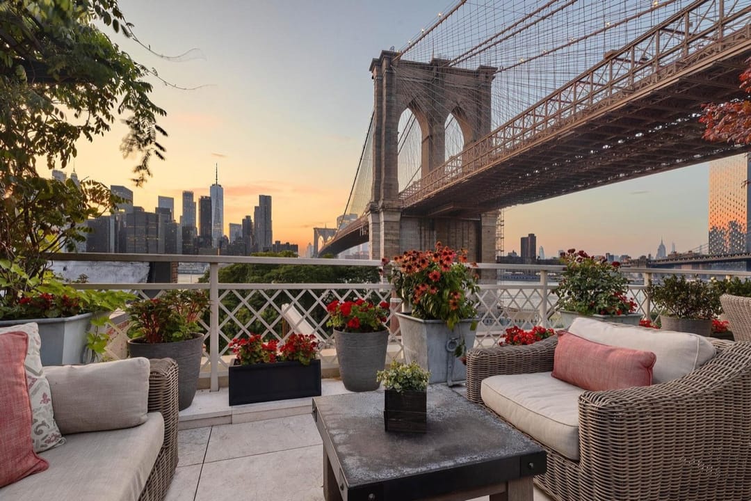 Terrace with wicker sofas and red flowers below the Brooklyn Bridge, Manhattan skyline at sunset